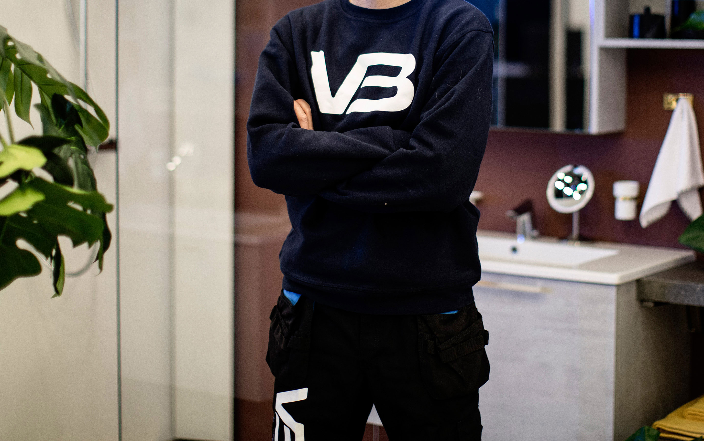 VB-rørlegger med arbeidsgenser med VB-logo, armene i kors foran servant og speilskap på burgunder vegg.